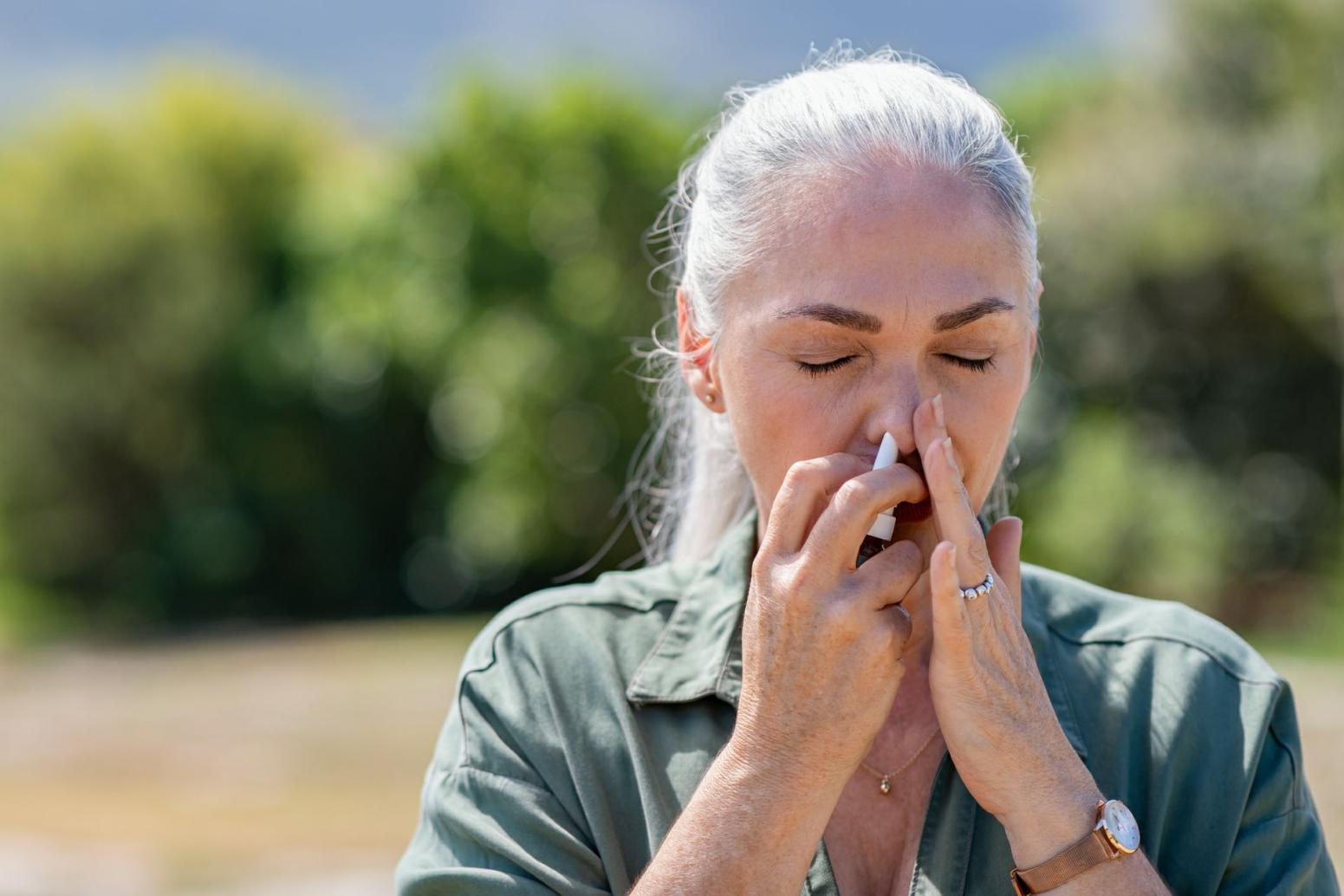 Woman with grey hair closes left nostril with hand and uses white nasal spray bottle in right nostril